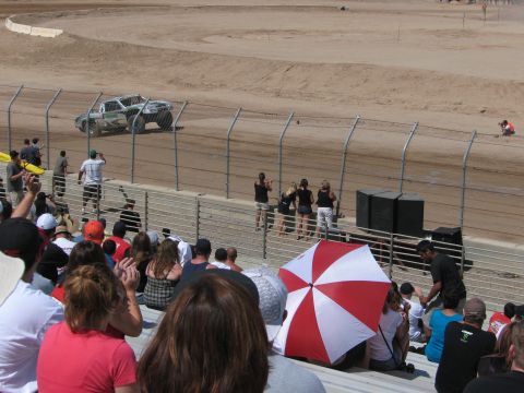 1990 Ford Bronco - white off-road race truck rounds a dirt corner while a large crowd, including a red-and-white umbrella, watches from the grandstand.