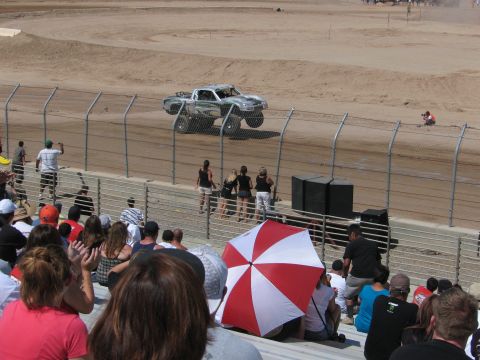 1990 Ford Bronco - white off-road race truck climbs a banked dirt section as fans crowd the grandstand and a red-and-white umbrella stands out in front.