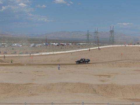 1990 Ford Bronco - dark pickup-style race truck drives a wide dirt course across an open desert valley lined with power transmission towers.