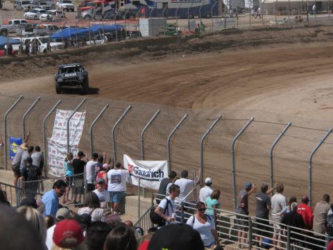 1990 Ford Bronco - dark off-road race truck kicks up dust on a banked dirt track as a crowd cheers near a BFGoodrich banner on the fence.