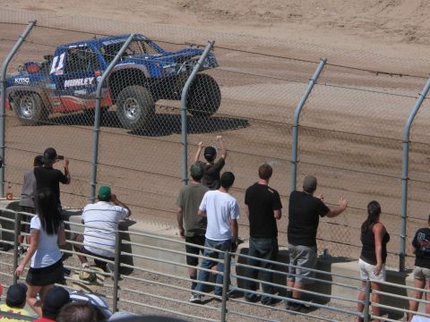 1990 Ford Bronco - blue number 11 off-road race truck rounds a dirt track as fans line the chain-link fence waving and taking photos.