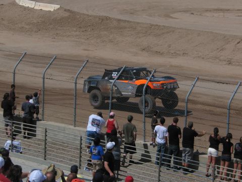 1990 Ford Bronco - orange and black number 22 off-road race truck drives a dirt track while spectators watch from the grandstand behind the fence.
