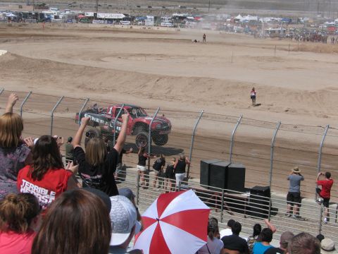 1990 Ford Bronco - red off-road race truck catches air on a dirt track as an animated crowd, one fan in a red shirt, cheers from the stands.
