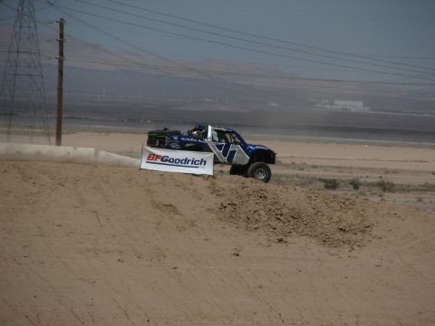 1990 Ford Bronco - blue number 7 off-road race truck goes airborne over a dirt jump near a BFGoodrich banner in a dusty desert setting.