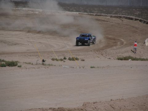 1990 Ford Bronco - blue off-road race truck slides through a dusty dirt corner trailing a cloud of dust, with a course marshal standing trackside.