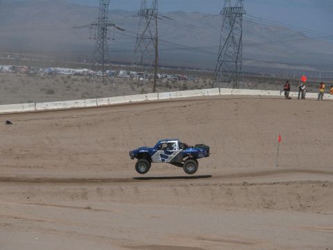 1990 Ford Bronco - blue and white off-road race truck launches off a jump on a banked dirt track beneath power transmission towers in the desert.