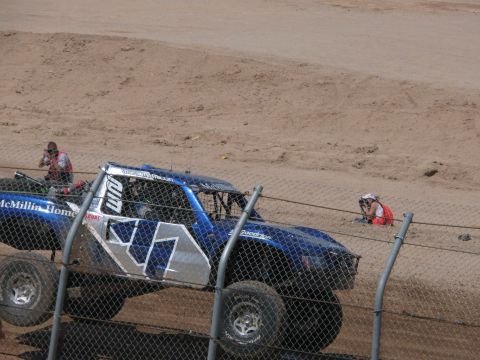 1990 Ford Bronco - blue off-road race truck marked McMillin Homes drives a dirt track seen through chain-link fencing, with crew members trackside.