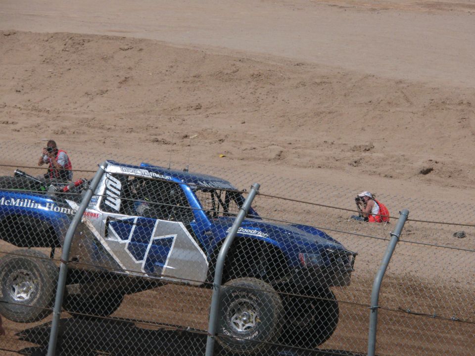 Blue McMillin Homes off-road race truck on a dirt track behind fencing