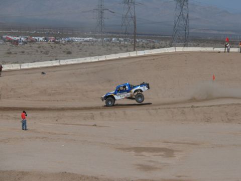 1990 Ford Bronco - blue and white off-road race truck powers around a banked dirt corner in the desert as a marshal watches trackside.