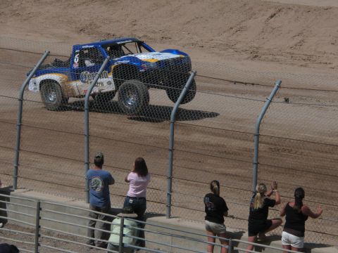 1990 Ford Bronco - blue off-road race truck with Coors branding drives a dirt track while fans watch and take photos along the chain-link fence.