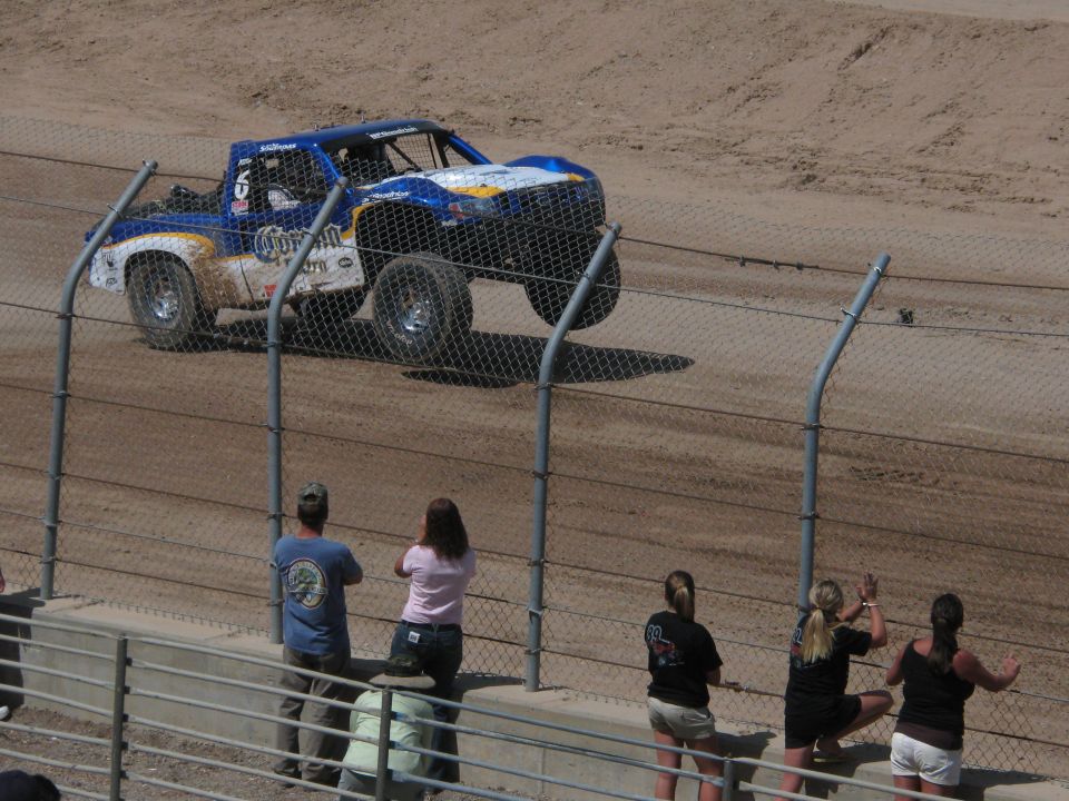 Blue Coors-branded off-road race truck on a dirt track with fans at fence