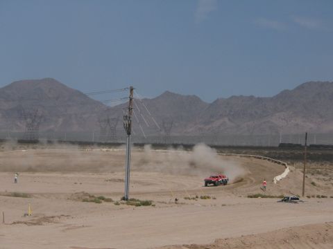 1990 Ford Bronco - red off-road race truck trails a plume of dust on a wide desert dirt course backed by transmission towers and distant mountains.