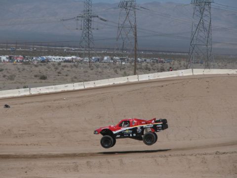 1990 Ford Bronco - red off-road race truck catches air on a banked dirt track beneath power transmission towers in a wide desert valley.