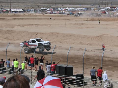 1990 Ford Bronco - white number 12 off-road race truck jumps a dirt rise as fans cheer along the fence at a desert short-course event.