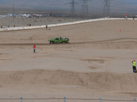1990 Ford Bronco - green off-road race truck drives a wide dirt desert course as course marshals stand trackside beneath transmission towers.