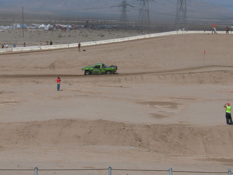 Green off-road race truck on a wide dirt desert course with marshals