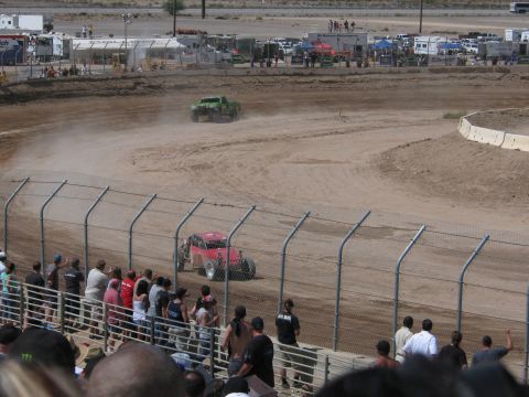 1990 Ford Bronco - red off-road race truck chases a green one through a dusty dirt corner as spectators watch from the grandstand fence.