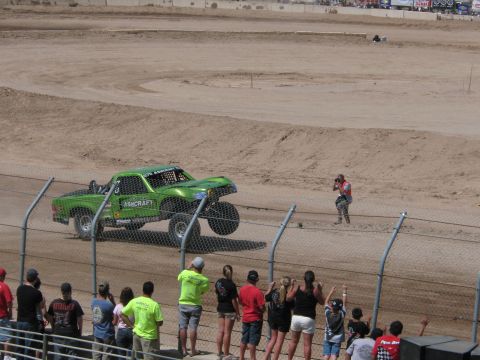 1990 Ford Bronco - green off-road race truck marked Ashcraft drives a dirt track while fans in yellow and other shirts watch along the fence.