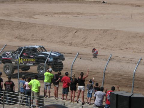 1990 Ford Bronco - black number 23 off-road race truck with Rockstar Energy sponsorship speeds along a dirt course as fans watch from behind a chain-link fenc…