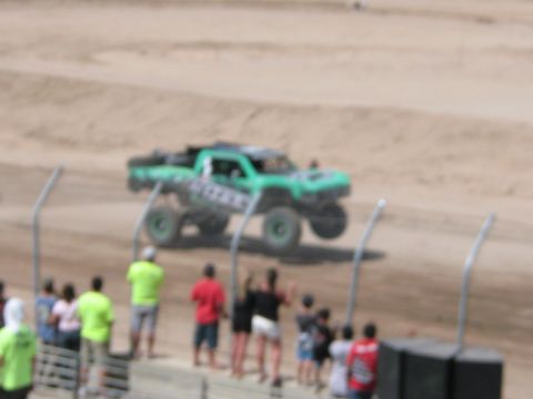 1990 Ford Bronco - green off-road race truck catches air over a dirt jump in a motion-blurred shot as spectators watch from the stands.
