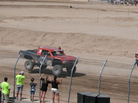 1990 Ford Bronco - red and black off-road race truck rounds a dirt track as a small group of spectators watches from behind a chain-link fence.