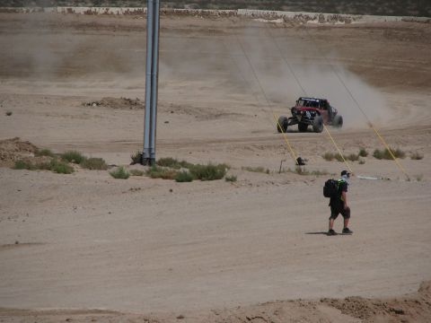 1990 Ford Bronco - off-road race buggy speeds across a dusty desert course while a person carrying gear walks across the dirt in the foreground.