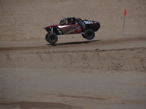 1990 Ford Bronco - black and red off-road race truck launches off a dirt rise near an orange course-marker flag in the desert.