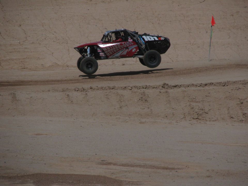Black and red off-road race truck airborne over dirt with orange course flag