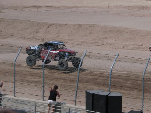 1990 Ford Bronco - black and red off-road race truck drives along a dirt track behind a chain-link fence with a few spectators nearby.
