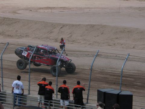 1990 Ford Bronco - red off-road race truck rounds a dirt course as a crew in black and orange team jerseys watches from behind the fence.