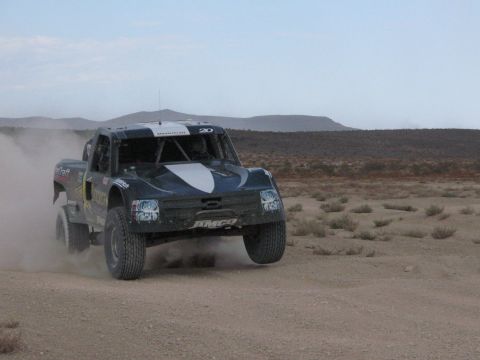 1990 Ford Bronco - dark-colored number 20 off-road race truck kicks up dust as it races across open desert with hills in the background.