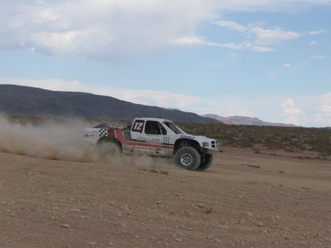 1990 Ford Bronco - white and red number 12 off-road race truck speeds through the desert trailing a cloud of dust under a partly cloudy sky.