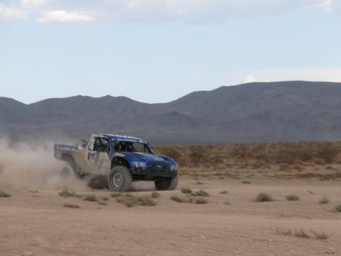 1990 Ford Bronco - blue off-road race truck races across a sandy desert flat, kicking up dust with mountains in the distance.
