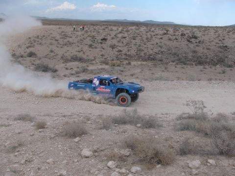 1990 Ford Bronco - blue number 24 off-road race truck slides through a dusty turn on a desert course with rocky scrub around it.