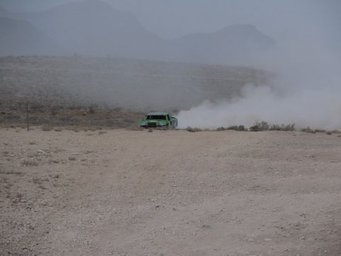 1990 Ford Bronco - green off-road race truck races into a thick dust cloud on a desert course with hazy mountains behind.
