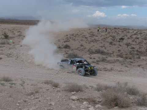 1990 Ford Bronco - off-road race truck trails a plume of dust through a desert turn as a few spectators watch from a ridge in the distance.