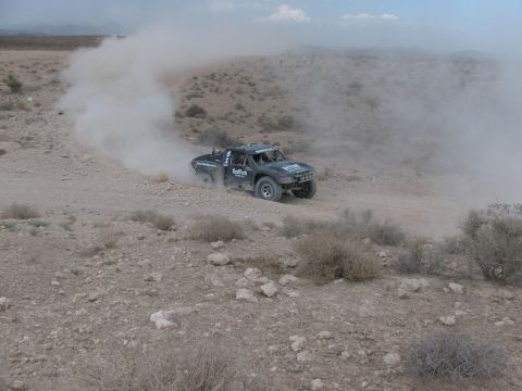 1990 Ford Bronco - dark off-road race truck slides through a dusty curve on a desert course bordered by rocks and scrub brush.