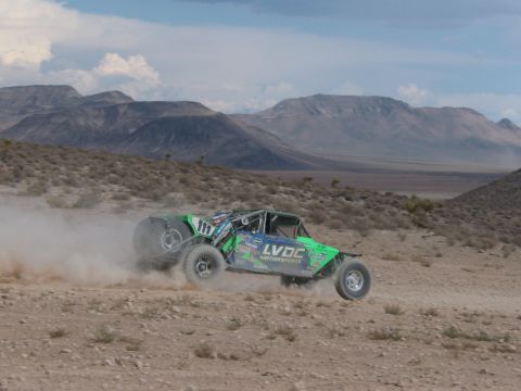 1990 Ford Bronco - green and blue number 117 off-road race truck with LVOC Motorsports livery races across the desert below distant mountains.