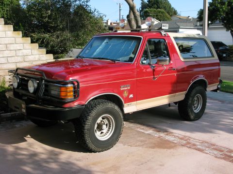 1989 Ford Bronco - A red 1987 Ford Bronco is parked on a driveway, equipped with auxiliary lights and off-road tires.