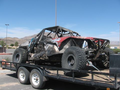 1990 Ford Bronco - fire-damaged tube-chassis off-road race car, its body burned away, sits strapped to a flatbed trailer in a parking lot.