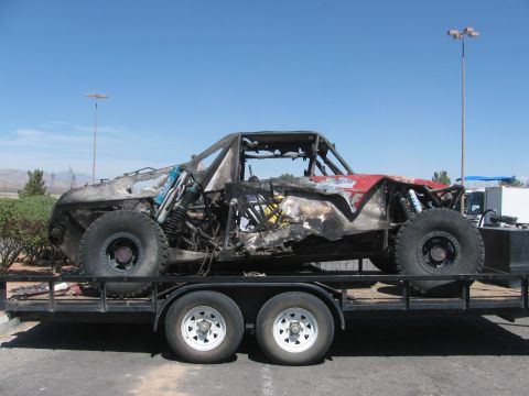 1990 Ford Bronco - side view of a fire-destroyed tube-frame off-road race car loaded on a flatbed trailer in a paved lot under clear skies.