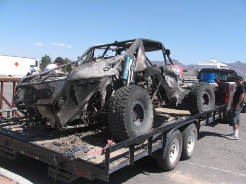 1990 Ford Bronco - charred tube-chassis off-road race car rests on a flatbed trailer while a man stands beside it in a parking lot.