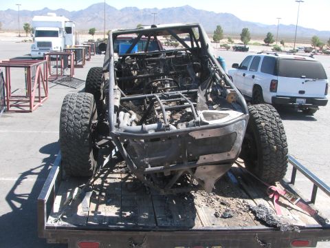 1990 Ford Bronco - rear of a burned-out off-road race car shows its scorched tube frame and bare engine bay while loaded on a flatbed trailer.