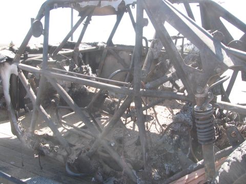 1990 Ford Bronco - close-up of a fire-destroyed off-road race car shows its melted, ash-coated tube cage, coil-over shock, and steering linkage.