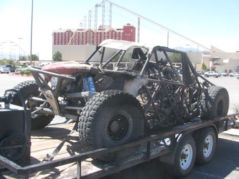 1990 Ford Bronco - burned tube-chassis off-road race car sits on a flatbed trailer with a large hotel and roller coaster visible in the background.