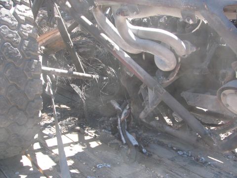 1990 Ford Bronco - close-up under a burned off-road race car shows scorched exhaust headers, a coil-over shock, and an ash-covered tire.