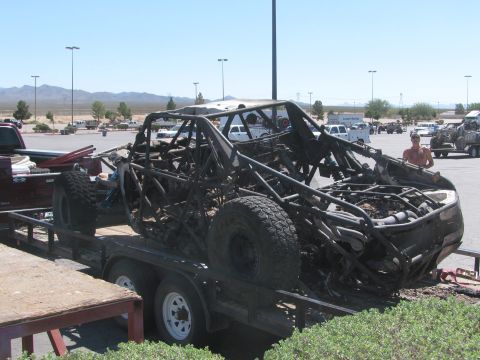 1990 Ford Bronco - burned-out off-road race car sits on a flatbed trailer in a desert parking lot, with a man and other vehicles in the background.