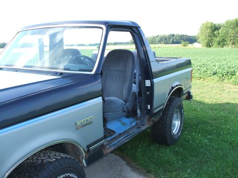 1990 Ford Bronco - A two-tone Ford Bronco is parked on grass with an open driver's door, revealing its interior and a field in the background.