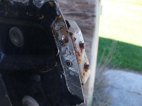 1990 Ford Bronco - A heavily rusted metal bracket with visible bolts is attached to a dark surface, with a wooden post and grass in the background.