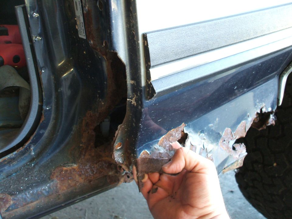 Close-up of severe rust on a vehicle's door frame and rocker panel, with a hand touching a flaking piece
